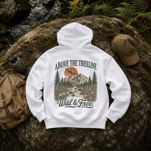 A white pullover hoodie laid flat on a mossy rock outdoors, showing the back graphic with the text 'ABOVE THE TREELINE' and 'Wild & Free' around a mountain, trees, and sunset illustration. A tan cap, backpack, and water bottle are placed around it in a forest setting.