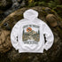 A white pullover hoodie laid flat on a mossy rock outdoors, showing the back graphic with the text 'ABOVE THE TREELINE' and 'Wild & Free' around a mountain, trees, and sunset illustration. A tan cap, backpack, and water bottle are placed around it in a forest setting.