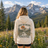 A woman stands outdoors in a mountain meadow with pine trees and rocky peaks in the background, viewed from behind. She is wearing a cream hoodie with a large back graphic that reads “ABOVE THE TREELINE” and “Wild & Free,” featuring an illustrated mountain, trees, and sunset.