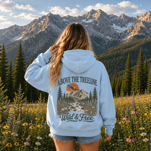 A woman stands in a mountain meadow with snow-capped peaks in the background, viewed from behind. She is wearing a light blue hoodie with a large back graphic that reads “ABOVE THE TREELINE” and “Wild & Free,” plus an illustrated mountain, trees, river, and bird scene.