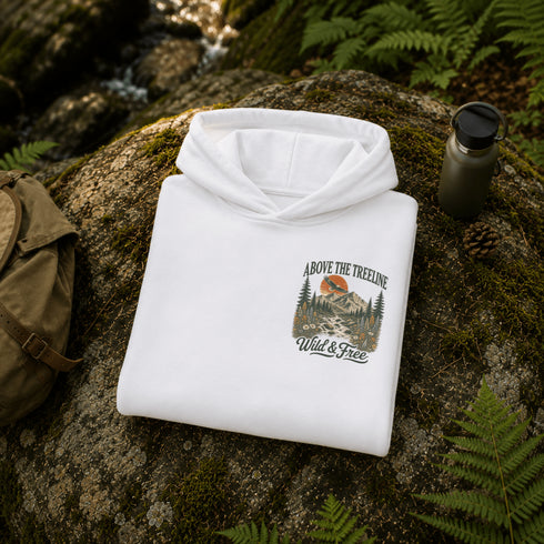 A folded white hoodie is placed on a mossy rock outdoors, with ferns, a backpack, and a water bottle nearby. The hoodie has a front graphic reading 'ABOVE THE TREeline Wild & Free' with a mountain-and-trees illustration.