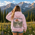 A woman stands in a mountain meadow with pine trees and snow-capped peaks in the background, wearing a light pink hoodie from the back. The hoodie features a large graphic print with the text 'ABOVE THE TREeline' and 'Wild & Free' surrounding a mountain, tree, and sunset scene.
