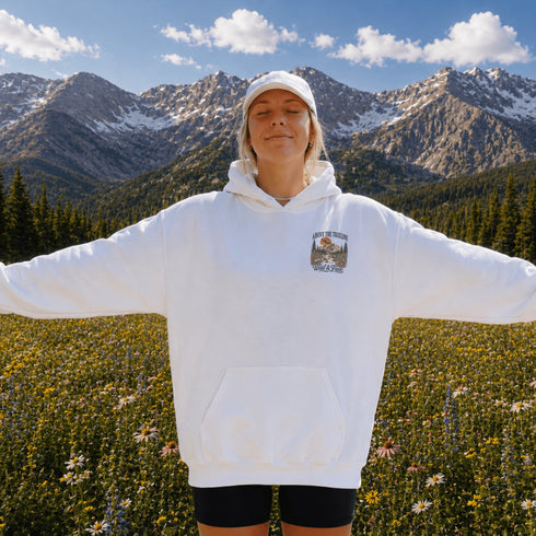 A woman stands in a mountain meadow with her arms outstretched, wearing a white pullover hoodie with a small mountain graphic on the left chest and a white cap. Snow-capped mountains and a blue sky are visible in the background.