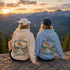 Two people sitting on a rocky mountain overlook at sunset, seen from behind, wearing matching hoodies with a large forest and mountain graphic and the text 'ABOVE THE TREeline' and 'Wild & Free' on the back.