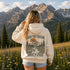 A woman stands in a mountain meadow with pine trees and rugged peaks in the background, viewed from behind. She is wearing a cream hoodie with a large back graphic that reads “ABOVE THE TREeline” and “Wild & Free,” plus a mountain, trees, river, and bird illustration.