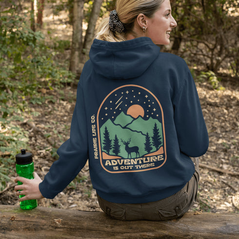 This graphic hoodie features a majestic deer standing against a wilderness backdrop— mountains, towering pines, a glowing moon, and a star-filled night sky.