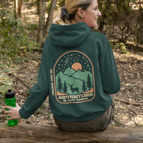 This graphic hoodie features a majestic deer standing against a wilderness backdrop— mountains, towering pines, a glowing moon, and a star-filled night sky.