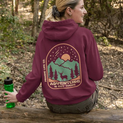 This graphic hoodie features a majestic deer standing against a wilderness backdrop— mountains, towering pines, a glowing moon, and a star-filled night sky.