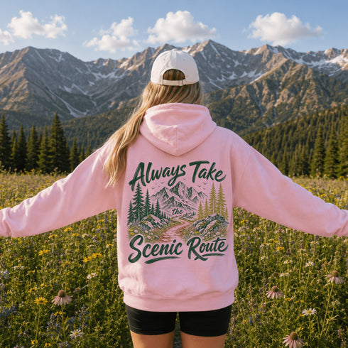 A woman stands in a mountain meadow with her back to the camera, wearing a light pink hoodie with a large graphic on the back that reads 'Always Take Scenic Route' above and below a pine forest and mountain illustration. She is also wearing a white baseball cap and black shorts, with rugged mountains and evergreen trees in the background.