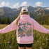 A woman stands in a mountain meadow with her back to the camera, wearing a light pink hoodie with a large graphic on the back that reads 'Always Take Scenic Route' above and below a pine forest and mountain illustration. She is also wearing a white baseball cap and black shorts, with rugged mountains and evergreen trees in the background.