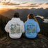 Two people sitting on a rocky overlook at sunset, seen from behind, wearing matching hoodies with a scenic mountain and lake graphic and the words 'Always Take Scenic Route.'.