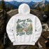 A white hooded sweatshirt laid flat on a rock with a mountain-and-forest landscape printed on the back. The graphic reads “Always Take the Scenic Route” in dark green script over an illustrated trail, trees, and mountains.