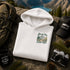 A folded white hoodie with a green and yellow scenic mountain graphic and the words 'Always Take Scenic Route' is displayed on a rock outdoors, surrounded by camping props including an olive green backpack, a black 'Adventure Awaits' mug, a black 'Explore More' cap, a vintage camera, and pine needles.