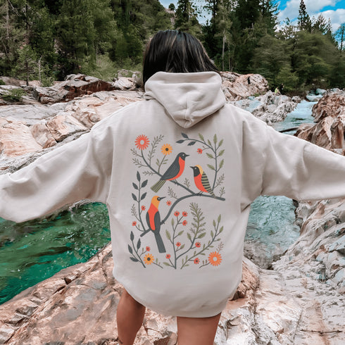 Wrap yourself in the charm of nature with our Among the Leaves Hoodie, a wearable piece of folk art that celebrates the harmony of birds, branches, and blooms.