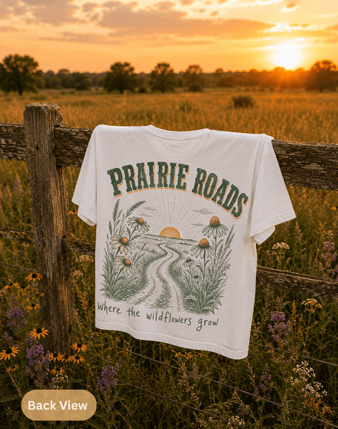 T-shirt with 'Prairie Roads' design hanging on a wooden fence in a field at sunset.