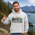 Man wearing a hoodie with a British Columbia design, pointing to it, with a scenic background of mountains and water.