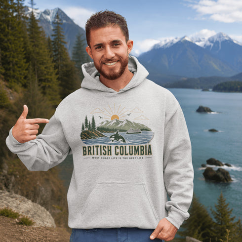 Man wearing a hoodie with 'British Columbia' design, pointing to it, with a scenic background of mountains and water.
