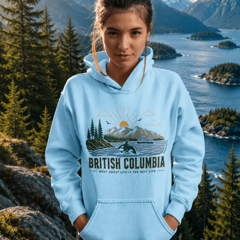 Person wearing a light blue hoodie with a 'British Columbia' design, standing in front of a scenic mountain and lake backdrop.