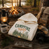 A cream-colored hooded sweatshirt with a 'CAMPFIRE nights' graphic is displayed in a rustic campfire setting on a wooden table, surrounded by a lantern, coffee mug, camera, and backpack.