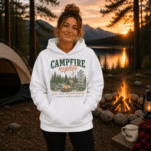 A woman stands outdoors at a campsite wearing a white pullover hoodie with a large 'CAMPFIRE nights' graphic on the front. The background shows a lake, mountains, sunset, a tent, a campfire, and a mug.
