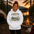 A woman stands outdoors at a campsite wearing a white pullover hoodie with a large 'CAMPFIRE nights' graphic on the front. The background shows a lake, mountains, sunset, a tent, a campfire, and a mug.