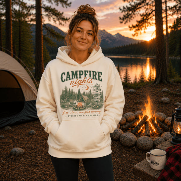 A woman stands outdoors at a campsite at sunset wearing a cream-colored hoodie with a 'Campfire Nights' forest graphic. A tent, a campfire, and a lake are visible in the background.