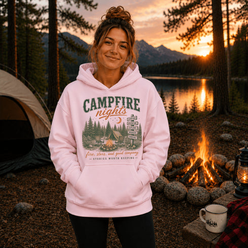 A woman stands outdoors at a campsite wearing a light pink hoodie with a rustic forest-themed graphic that reads 'CAMPFIRE nights' and 'fore stars and gold company - KANSAS NORTH WISCONSIN.' A tent, campfire, mug, and lantern are visible in the background at sunset.