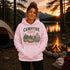 A woman stands outdoors at a campsite wearing a light pink hoodie with a rustic forest-themed graphic that reads 'CAMPFIRE nights' and 'fore stars and gold company - KANSAS NORTH WISCONSIN.' A tent, campfire, mug, and lantern are visible in the background at sunset.