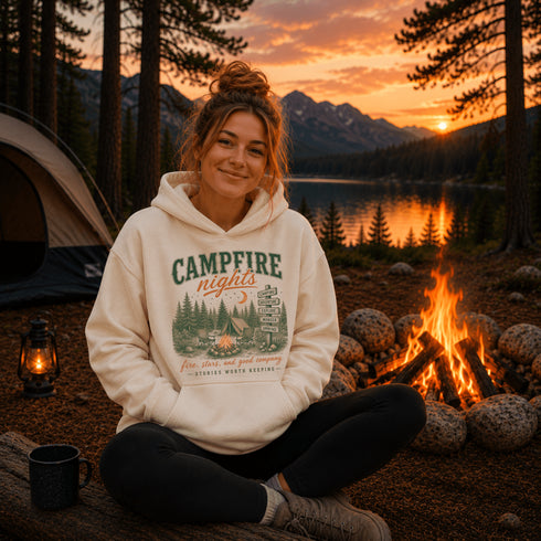 A woman sits cross-legged at a campfire beside a lake and mountains at sunset, wearing a cream hoodie with a green and orange 'Campfire Nights' graphic print.