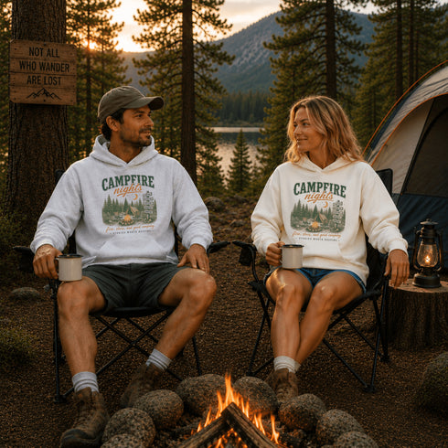 A man and a woman sit in folding chairs by a campfire at a lakeside campsite, wearing matching white hoodies with a 'CAMPFIRE nights' graphic print. A tent, trees, and a sunset are visible in the background.