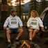A man and a woman sit in folding chairs by a campfire at a lakeside campsite, wearing matching white hoodies with a 'CAMPFIRE nights' graphic print. A tent, trees, and a sunset are visible in the background.