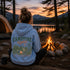 A person sits with their back to the camera at a lakeside campsite at sunset, wearing a light blue hoodie with a large green and orange 'CAMPFIRE nights' graphic on the back. A campfire burns in a stone ring nearby, with a tent and pine trees in the background.