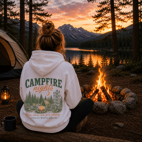 A woman sits at a campfire at a lakeside campsite at sunset, viewed from behind. She wears a white hoodie with a large back graphic reading “CAMPFIRE nights” and an illustrated camp scene.