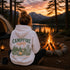 A woman sits at a campfire at a lakeside campsite at sunset, viewed from behind. She wears a white hoodie with a large back graphic reading “CAMPFIRE nights” and an illustrated camp scene.