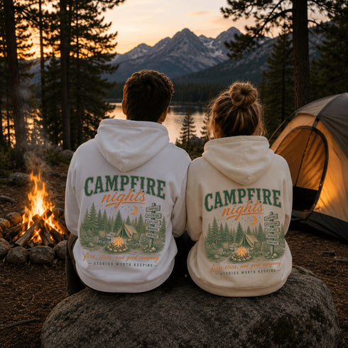 Two people sitting on a rock at a campsite, seen from behind, wearing matching white hoodies with a green and orange 'CAMPFIRE nights' graphic on the back. A campfire burns to the left, a tent is on the right, and mountains and a lake are visible in the background at sunset.