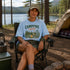 Person sitting in a campsite wearing a 'Campfire Nights' t-shirt with a scenic background.