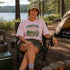 Person sitting in a campsite wearing a 'Campfire Nights' t-shirt with a scenic background.