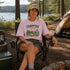 Person sitting in a campsite wearing a 'Campfire Nights' t-shirt with a scenic background.
