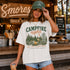 Woman wearing a 'Campfire Nights' t-shirt and green cap in front of a wooden snack bar with 'S'mores' sign.