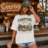 Woman wearing a 'Campfire Nights' t-shirt and green cap in front of a wooden snack bar with 'S'mores' sign.