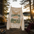 A cream-colored hoodie hanging on a clothesline at a lakeside campsite, featuring a green and orange graphic that reads 'CAMPFIRE nights' with a campfire, tents, and forest scene.