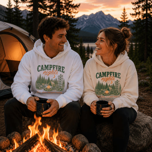 A man and a woman sitting by a campfire at dusk wearing matching white hoodies with a green and orange 'CAMPFIRE nights' graphic, with a tent, lake, and mountains in the background.
