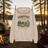 An off-white hooded sweatshirt hanging on a rope at a lakeside campsite, with a forest, tent, fire, mug, and lantern in the background. The hoodie has a large front graphic reading “CAMPFIRE nights” with trees, a campfire, and a signpost design.