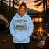 A woman stands outdoors at a campsite wearing a light blue hoodie with a 'CAMPFIRE nights' graphic print. The background features a campfire, a tent, a lake, and mountains at sunset.