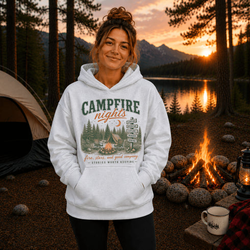 A woman stands outdoors at a campsite at sunset wearing a white pullover hoodie with a large vintage-style camp graphic that reads 'CAMPFIRE nights' and 'Seven Stars and Good Company - Outside North Dakota.' A tent, campfire, lake, mountains, and trees are visible in the background.