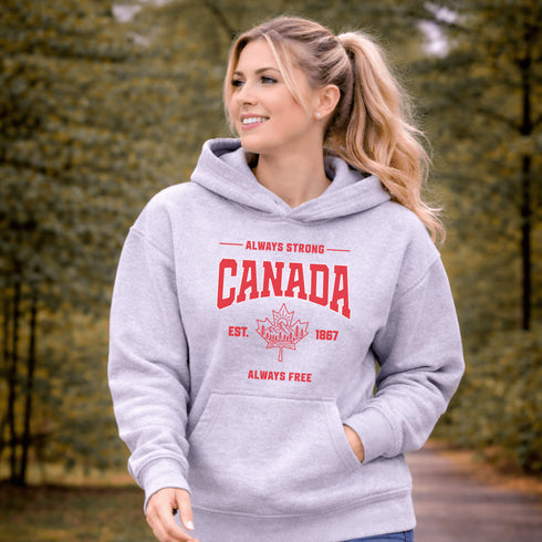 Woman wearing grey Canada Always Strong hoodie with red maple leaf graphic and “Est. 1867” design, styled outdoors in a natural setting