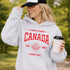Woman wearing light grey Canada Always Strong hoodie with red maple leaf graphic and “Est. 1867” design, styled outdoors in a natural setting