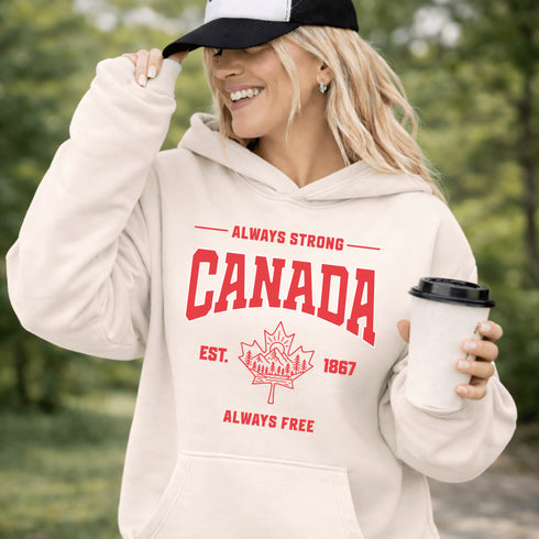Woman wearing white Canada Always Strong hoodie with red maple leaf graphic and “Est. 1867” design, styled outdoors in a natural setting