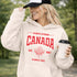 Woman wearing white Canada Always Strong hoodie with red maple leaf graphic and “Est. 1867” design, styled outdoors in a natural setting
