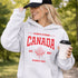 Woman wearing grey Canada Always Strong hoodie with red maple leaf graphic and “Est. 1867” design, styled outdoors in a natural setting